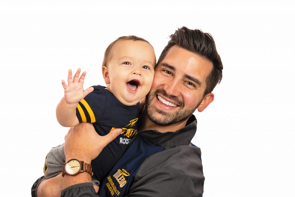 Father and Son smiling and laughing. Baby is waving. Healthy, bright smile. University of Toledo representative. University of Toledo gear. 